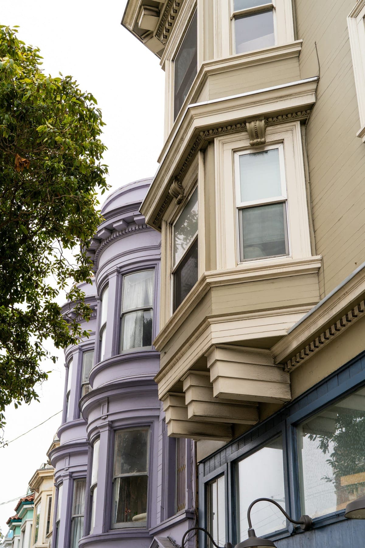 Stacked bay windows on a row of painted wooden flats, one cream and one lavender, beside a street tree.