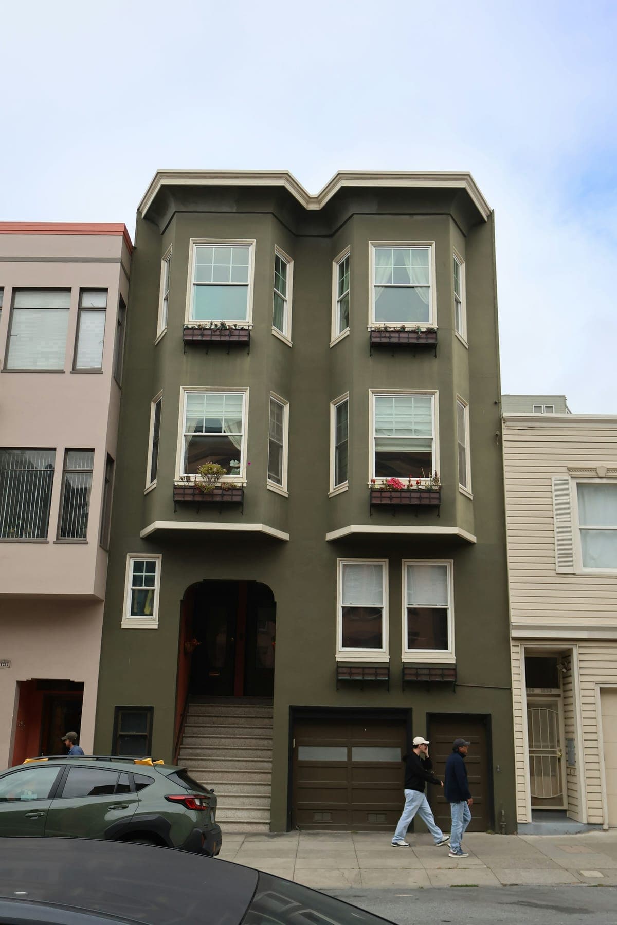 A three-storey olive green apartment building with two columns of bay windows, flower boxes and a garage at street level.