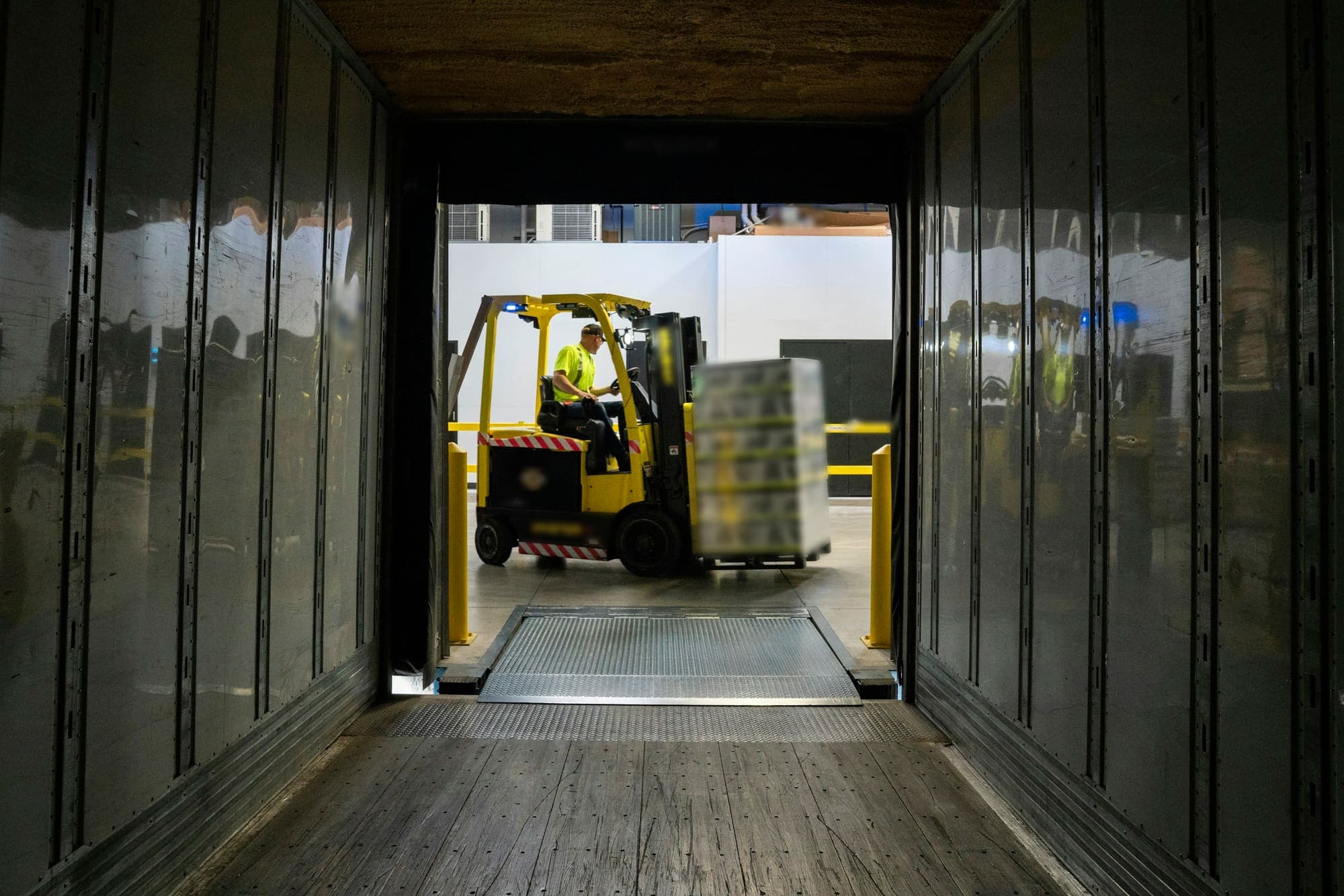 View from inside an empty trailer towards a loading dock, where a forklift carries a wrapped pallet to the dock plate