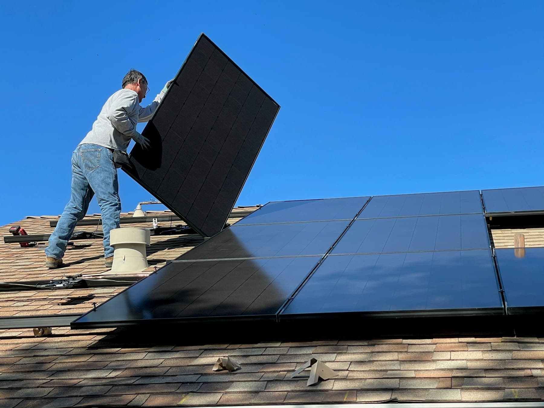 An installer carrying a black solar panel up a shingle roof under a deep blue sky
