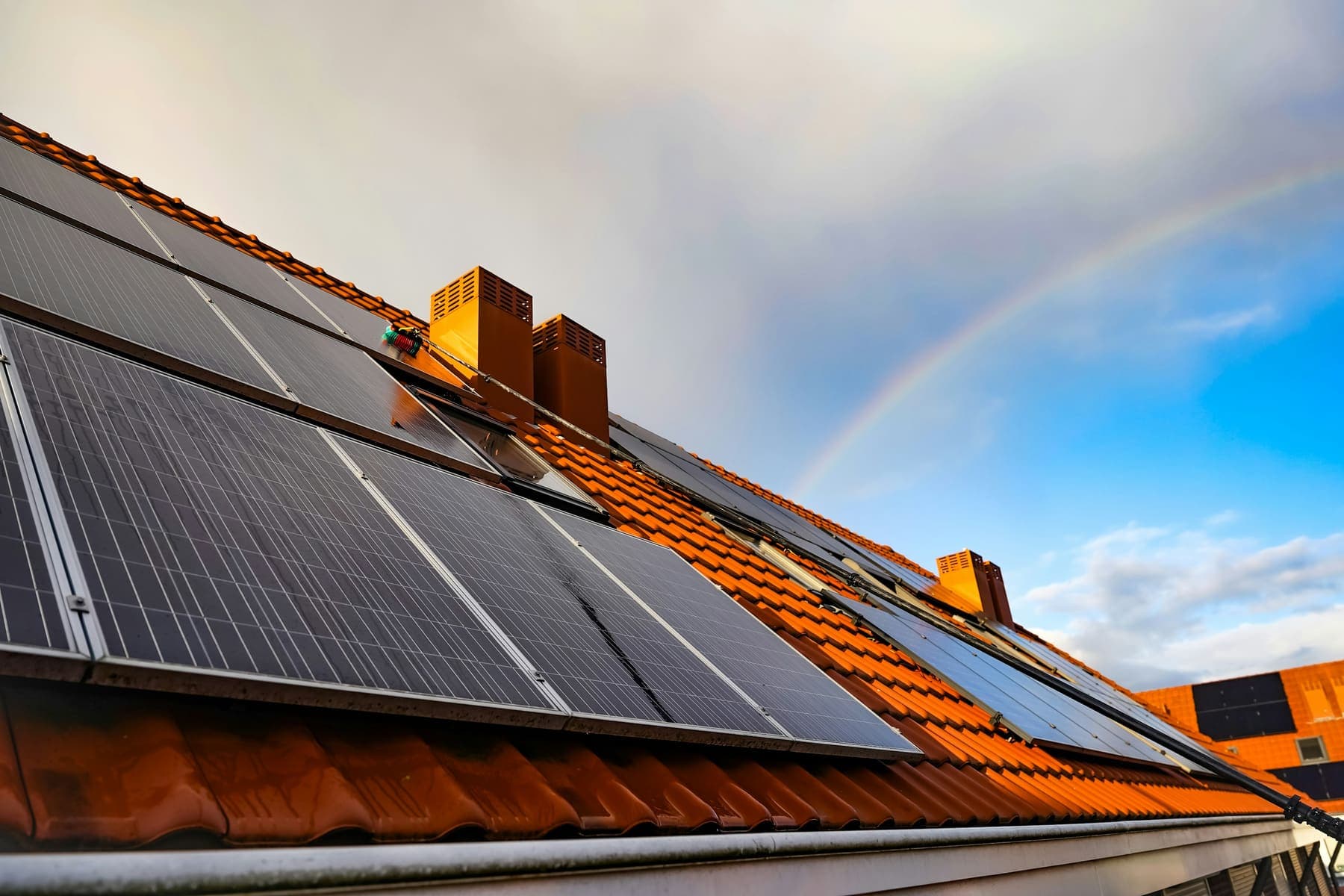 Rows of solar panels on a terracotta tile roof, a rainbow over clearing cloud behind